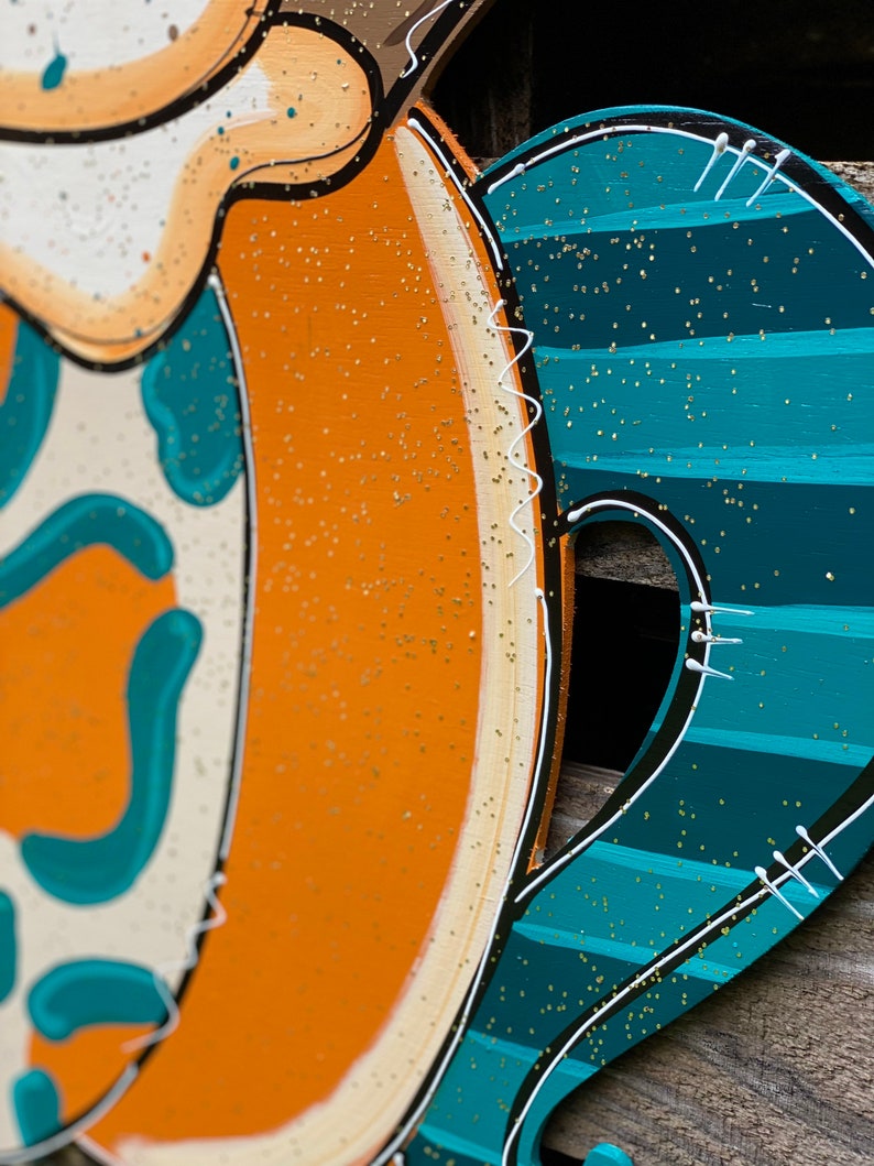 May include: A wooden door hanger shaped like a pumpkin with a teal and white striped design. The pumpkin is decorated with gold glitter and white stitching.