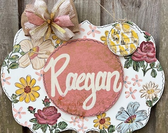 May include: A white wooden sign with a pink floral design and a pink circle with the name "Raegan" written in white. The sign also has a smaller yellow circle with the words "date, time, lbs, oz, in" written in black.