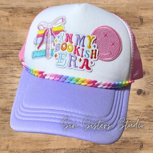 May include: A white and lavender trucker hat featuring a stack of colorful books with a bow, a pink smiley face patch, and the embroidered text IN MY BOOKISH ERA. The hat is accented with a rainbow-colored plastic chain trim along the brim base.