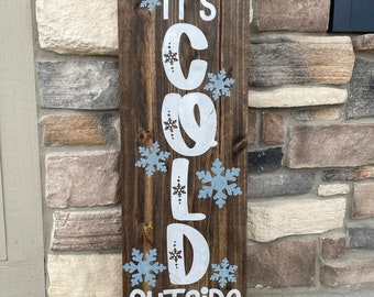 Può includere: Un alto cartello rettangolare in legno marrone scuro con la scritta "BABY IT'S COLD OUTSIDE" dipinta in bianco e disposta verticalmente. Il design è decorato con diverse illustrazioni di fiocchi di neve azzurri e bianchi.
