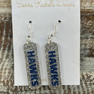 May include: Silver glitter earrings with blue 'HAWKS' lettering. The earrings are on a white card with the text 'Sienna Rachelle Designs'.