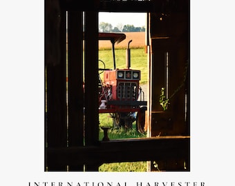 May include: A red International Harvester tractor viewed through a dark wooden barn doorway, featuring a prominent vertical exhaust pipe, a front grille, and headlights, with a field visible in the background. Text reads: INTERNATIONAL HARVESTER.