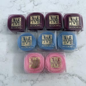 May include: An assortment of Midori Inc. thread spools in clear plastic boxes. The spools are in shades of purple, blue, and pink. Each box has a gold label with the letter "M" and the brand name.