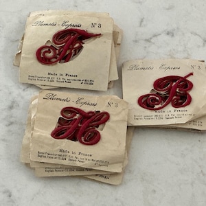May include: Three stacks of vintage Plumetis-Express No. 3 iron-on monograms in deep red thread. Each package features the letter F, B, or H in a cursive script, printed on cream-colored paper with the text Made in France and patent details.