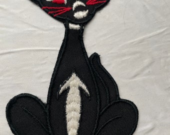 Op de afbeelding: Zwarte vilt kattenapplicatie met rode ogen en witte snorharen. De kat zit met zijn staart om zijn lichaam gekruld.