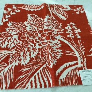 May include: A square fabric swatch featuring a bold orange and white botanical print with large leaves and berries. A white rectangular label in the bottom right corner displays the text Manuel Canovas Paris, Pali Lin, and technical specifications.