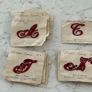 Op de afbeelding: Vier stapels vintage Plumetis Express nr. 3 strijkapplicaties met rode cursieve letters A, C, T en S. Elk papieren vierkant bevat de tekst Plumetis Express, nr. 3 en Made in France, samen met patentinformatie gedrukt in zwarte inkt.