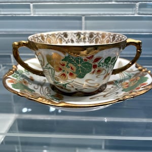 May include: A vintage teacup and saucer set. The white porcelain cup and saucer feature gold trim and floral designs in red, green, and gold. The cup has two gold handles. The saucer is scalloped with matching gold trim.