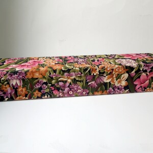 Puede incluir: Una caja larga y rectangular con un patrón floral. La caja está cubierta de una tela con fondo oscuro y flores de colores, incluyendo rosa, naranja, morado y verde.