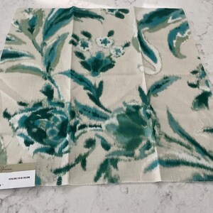 May include: A square fabric swatch featuring a floral pattern in shades of teal and sage green on a light beige background. A small label in the corner displays the text: Pattern: FLORA DANTE, Color: GREENTONE, Width: 55", Repeat: V 21.4" H 28.5".