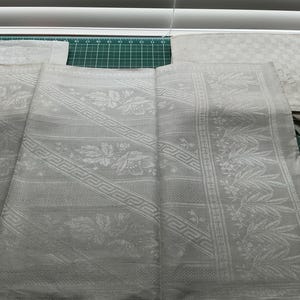 Può includere: Un tessuto grigio chiaro caratterizzato da un motivo damascato intrecciato con elementi floreali, bordi geometrici a greca e sezioni puntinate. Il materiale è piegato e disteso, mostrando un disegno decorativo ripetitivo sulla superficie.