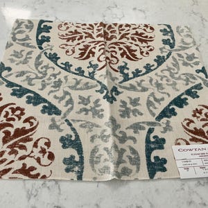 May include: A square fabric swatch featuring a cream background with a large-scale damask pattern in rust orange, teal, and grey. A white product label in the bottom right corner displays the text Cowtan & Tout, Amaranta, Sienna, Azure.