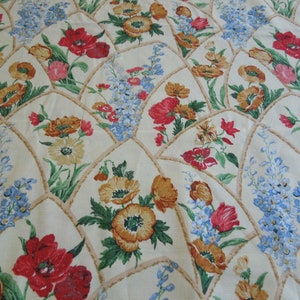 Op de afbeelding: Een crèmekleurige stof met een bloemenpatroon. Het patroon bevat rode klaprozen, gele bloemen en blauwe vergeet-me-nietjes. De bloemen zijn gerangschikt in een herhalend patroon binnen een geometrisch ontwerp.