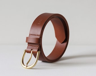 Peut inclure: Une ceinture en cuir marron avec une boucle dorée. La ceinture est enroulée et la boucle est face à l'appareil photo.