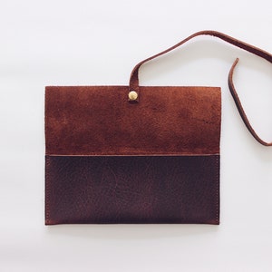 May include: A brown leather wallet with a flap closure and a strap. The wallet is open and the inside is visible. The wallet has a brown suede lining.