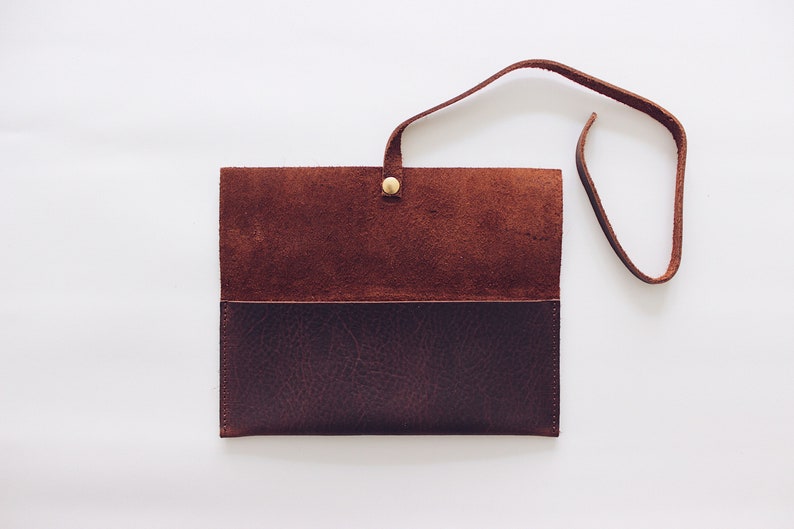May include: A brown leather wallet with a flap closure and a strap. The wallet is open and the inside is visible. The wallet has a brown suede lining.