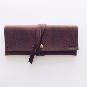 May include: A brown leather pencil case with a strap and a brass button closure. The case is personalized with the initials "JD" and the brand name "spady".