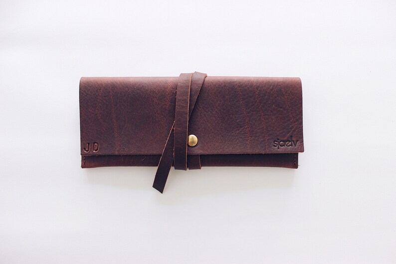 May include: A brown leather pencil case with a strap and a brass button closure. The case is personalized with the initials "JD" and the brand name "spady".