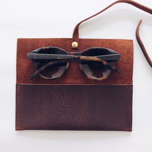 May include: A brown leather sunglasses case with a strap. The case is open and a pair of tortoiseshell sunglasses are inside.