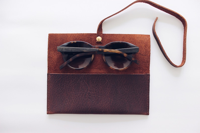 May include: A brown leather sunglasses case with a strap. The case is open and a pair of tortoiseshell sunglasses are inside.