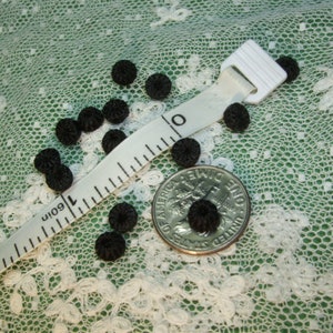 May include: Small, black, textured buttons scattered on a white lace surface. A white measuring tape shows inches, and a U.S. dime is included for size comparison. The buttons have a flower-like design.