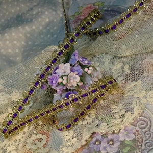 Puede incluir: Borde morado y dorado con borde festoneado. El borde está sobre un tejido de encaje blanco con un grupo de pequeñas flores moradas y blancas.