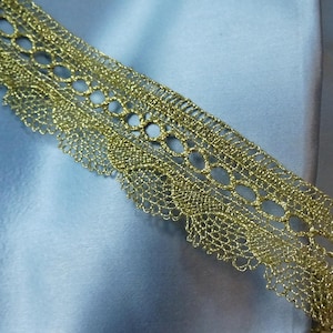 May include: Close-up of a gold-colored lace trim with an openwork design. The trim is made of a metallic thread and has a scalloped edge. The lace is set against a light blue fabric background, creating a contrast.