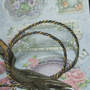 Op de afbeelding: Twee gedraaide touwarmbanden, één goud en zilver, de andere zilver en goud, worden getoond. De armbanden zijn geplaatst tegen een achtergrond van decoratief papier met bloemen- en vlinderthema's, met een zilveren zwaanbeeldje op de voorgrond.