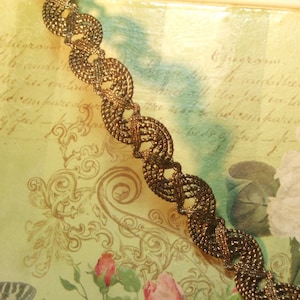 May include: Gold trim with a delicate, intricate design. The trim is woven in a circular pattern and has a slightly raised texture. The trim is on a background of a vintage-looking paper with faded writing.