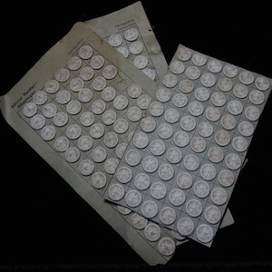 May include: Multiple sheets of small, circular, white objects with a starburst design are arranged on gray paper. The paper has a grid pattern and German text. The sheets are stacked on a black background.