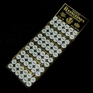 Op de afbeelding: Een kaart met een raster van witte knoppen met het label "Reinleinen Adlerknöpfe U 11 1/2" in het Duits. De kaart is goudkleurig met zwarte tekst en een klein adelaarlogo. De knoppen zijn gerangschikt in rijen en kolommen.