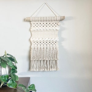 May include: A white macrame wall hanging with a wooden dowel at the top and fringe at the bottom. The wall hanging is made with a variety of knots and textures.