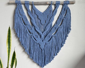 May include: A blue macrame wall hanging with a geometric design. The wall decor is suspended from a natural wooden dowel and features intricate knotting patterns and fringe. The piece is a decorative element for home interiors.