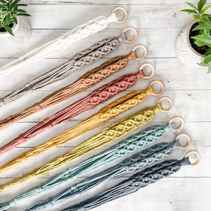 May include: A set of ten macrame plant hangers in various colors, including white, gray, brown, orange, yellow, green, and blue. Each hanger features a wooden ring at the top and a knotted design.
