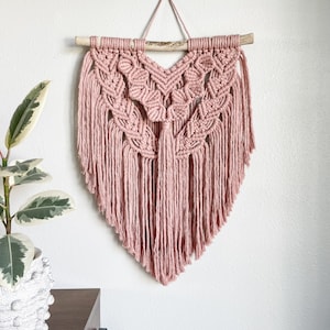 May include: A pink macrame wall hanging with a wooden dowel at the top and fringe details. The hanging is in the shape of a heart.