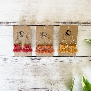 May include: Three pairs of handmade macrame earrings with triangle shapes and fringe. The earrings are displayed on a wooden surface with the Dakota Knot Company logo on each card.