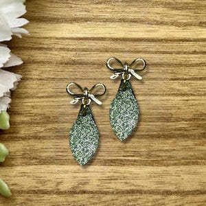 May include: A pair of dangle earrings featuring a gold-toned metal bow at the top and a teardrop-shaped pendant covered in fine green glitter. The earrings have a metallic finish and a textured, sparkling surface on the lower drop component.
