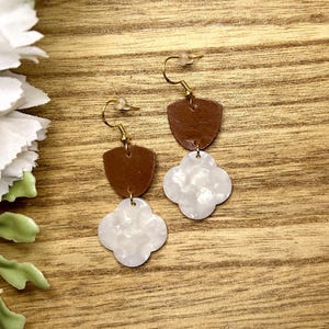 May include: A pair of drop earrings featuring a brown leather shield-shaped top piece connected to a white marbled acrylic clover-shaped pendant. The earrings are suspended from gold-toned metal fishhook wires with clear plastic backings.