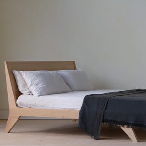 May include: A contemporary wooden bed frame with a low profile, in a light wood finish. The bed is made up with white linens and pillows, and a dark grey blanket with fringe. The bed is set against a neutral wall.