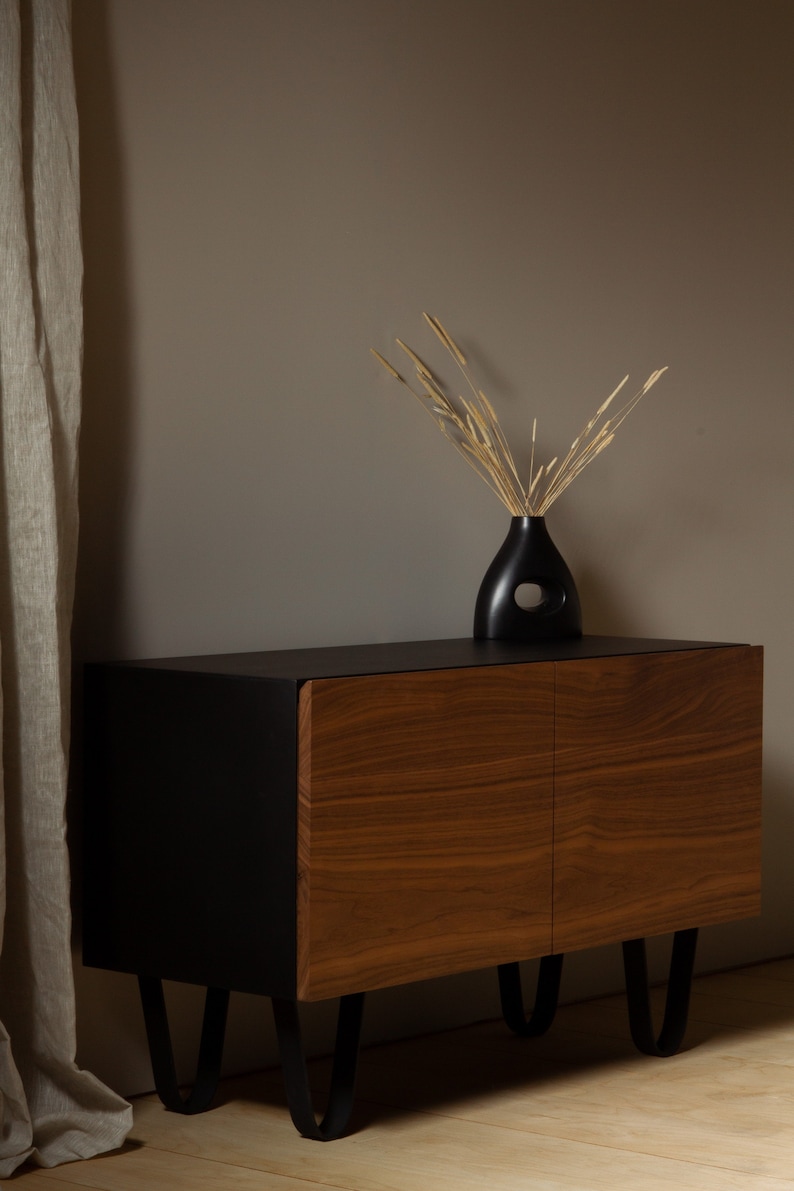 May include: A black cabinet with two wooden doors and black metal legs. A vase with dried flowers sits on top of the cabinet.