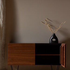 May include: A mid-century modern style wooden cabinet with black accents and hairpin legs. The cabinet has two doors, one of which is open, revealing an empty shelf. A black vase with a cutout design holds dried grasses.