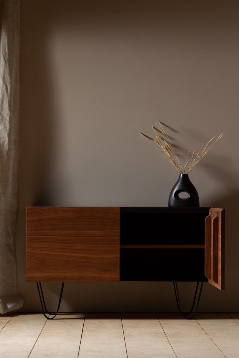 May include: A mid-century modern style wooden cabinet with black accents and hairpin legs. The cabinet has two doors, one of which is open, revealing an empty shelf. A black vase with a cutout design holds dried grasses.