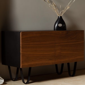 May include: A black and walnut wood cabinet with two doors and black metal hairpin legs. The cabinet is sitting on a light wood floor.