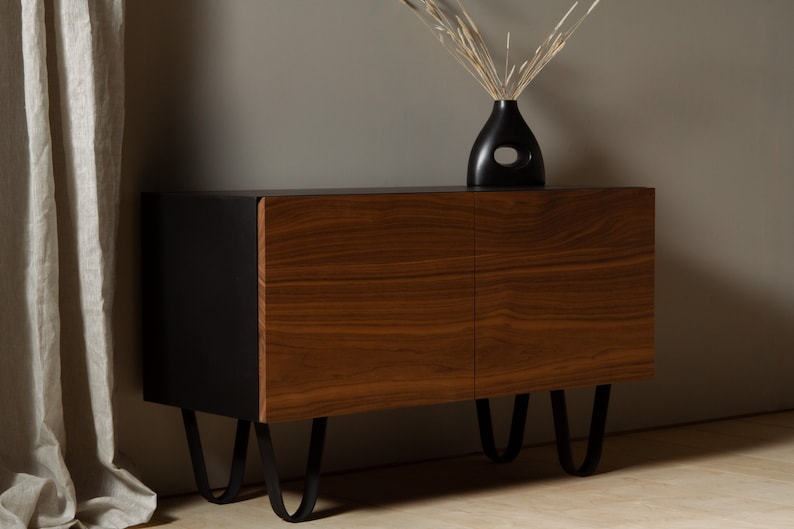 May include: A black and walnut wood cabinet with two doors and black metal hairpin legs. The cabinet is sitting on a light wood floor.