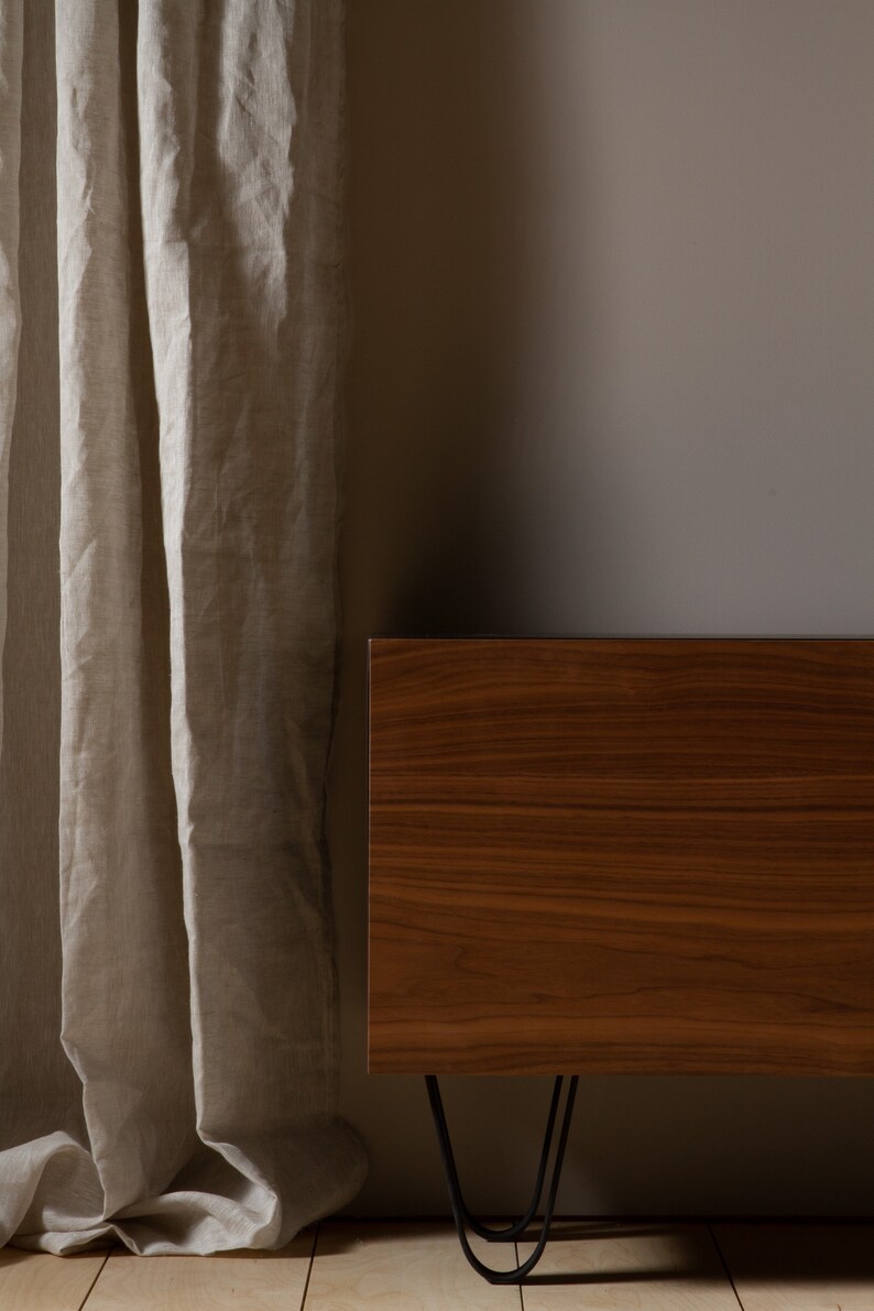 May include: A wooden cabinet with a dark brown finish and black hairpin legs. The cabinet is positioned in front of a light beige wall and a sheer linen curtain.