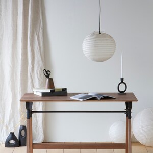 May include: A wooden desk with black metal supports and legs, topped with books, scissors, and a candle holder. A white paper lantern hangs above. Decorative vases and paper lanterns are on the floor.