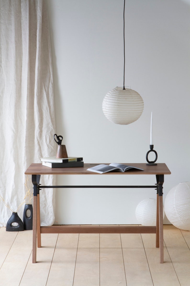 May include: A wooden desk with black metal supports and legs, topped with books, scissors, and a candle holder. A white paper lantern hangs above. Decorative vases and paper lanterns are on the floor.