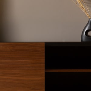 May include: A mid-century modern style cabinet with a wood veneer door and black shelves. The cabinet has black metal hairpin legs and is sitting on a light brown surface. A black vase with dried grasses is on the surface behind the cabinet.