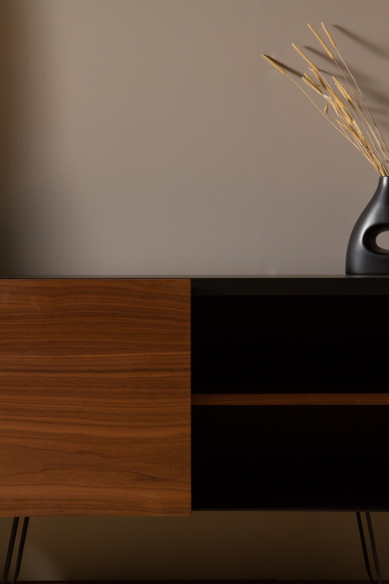 May include: A mid-century modern style cabinet with a wood veneer door and black shelves. The cabinet has black metal hairpin legs and is sitting on a light brown surface. A black vase with dried grasses is on the surface behind the cabinet.