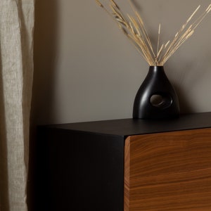 May include: A black wooden cabinet with a single drawer. The drawer is made of wood with a natural grain. The cabinet is sitting on a black surface. A black vase with a cutout design holds dried grasses.